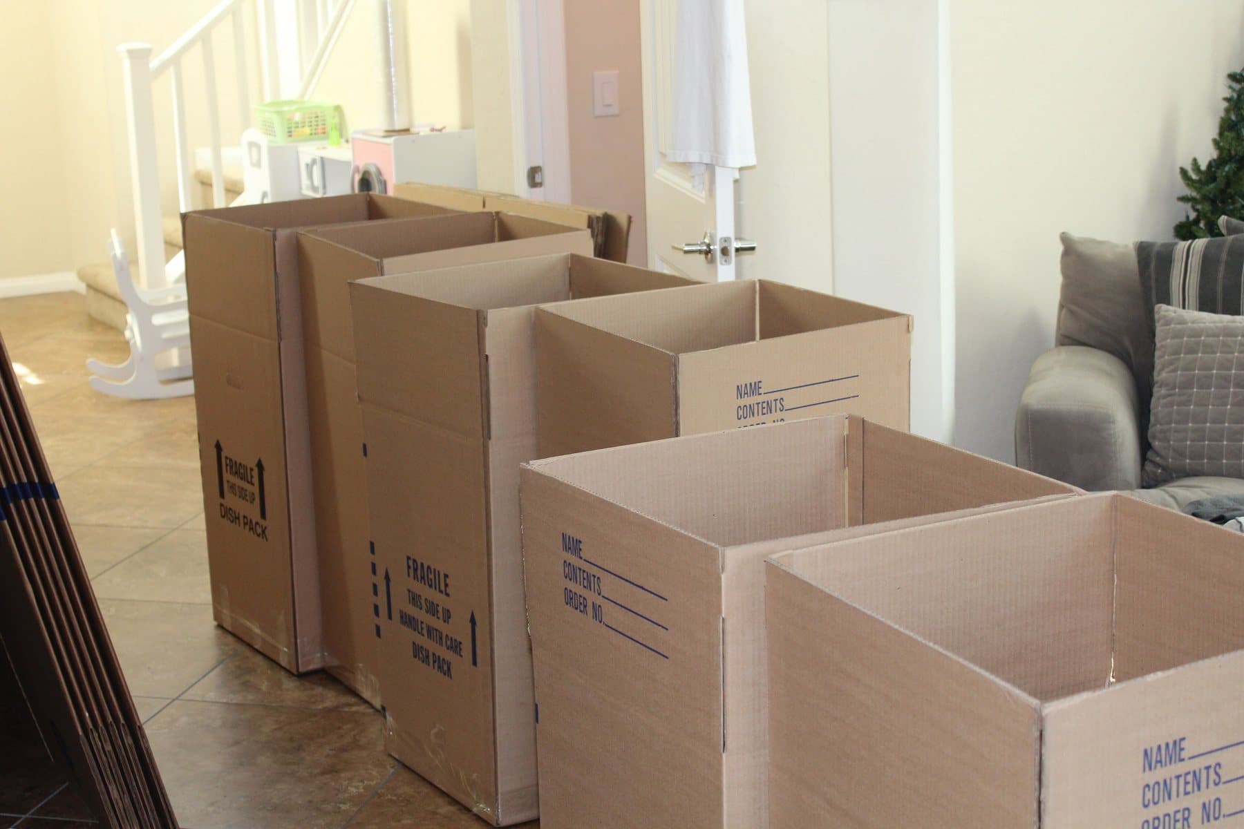A row of empty cardboard moving boxes, including tall dish-pack cartons, set out on a tiled floor in a bright living room